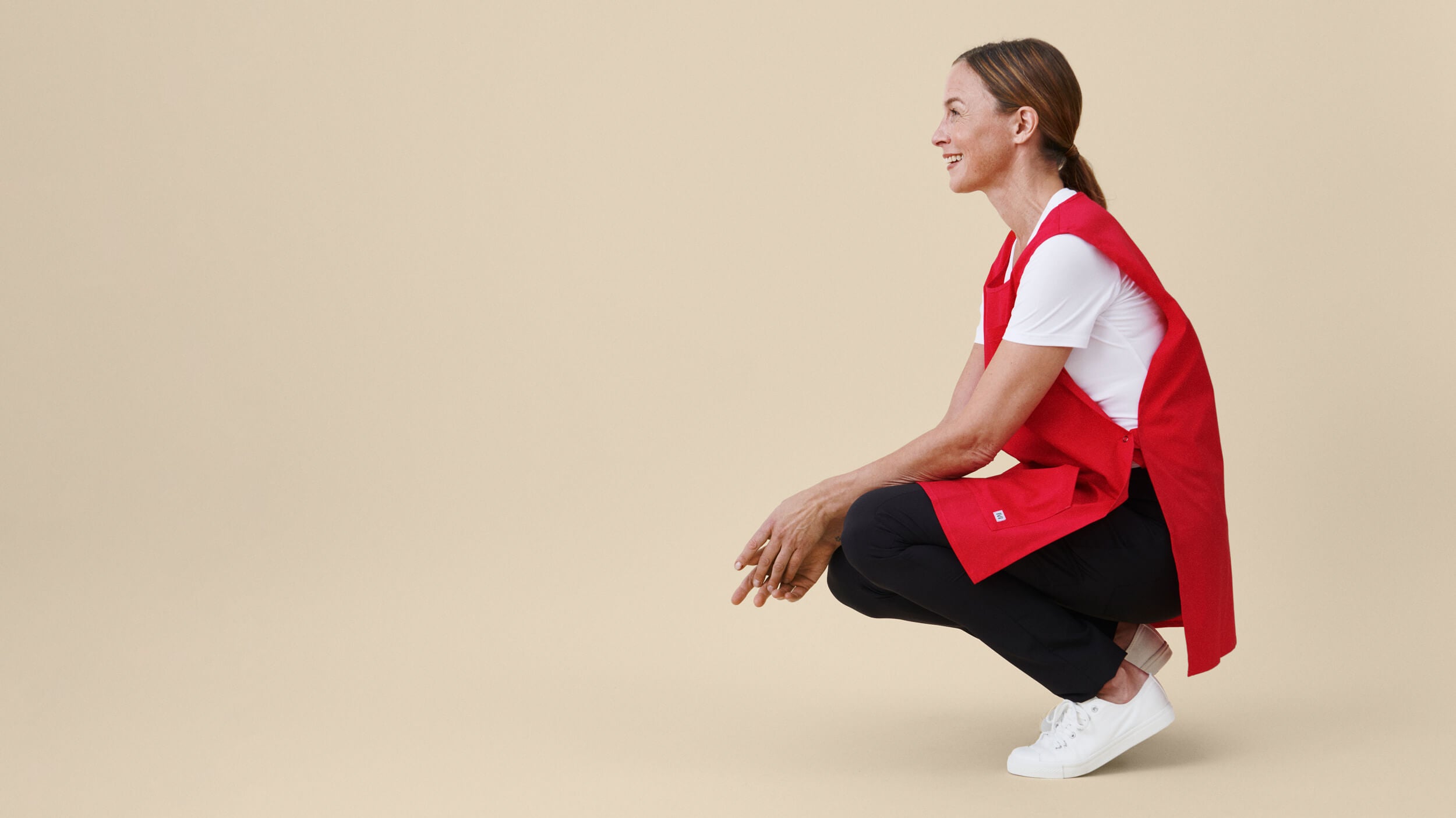 Punainen työessu, mustat työhousut ja valkoinen T-paita sivusuunnasta, hymyilevän hoitajan yllä. Beige tausta.