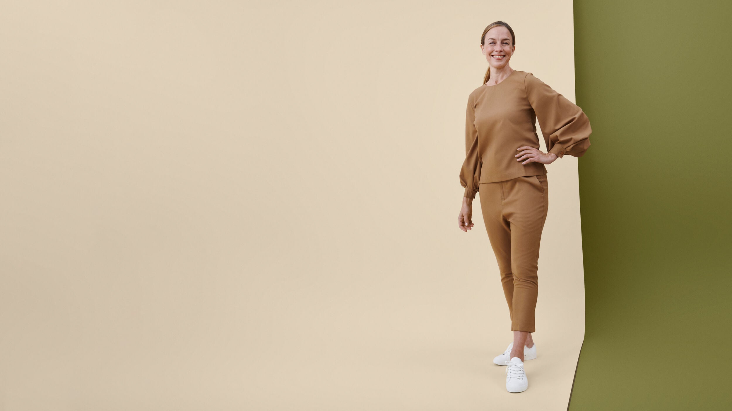 Vaaleanruskea työasu hymyilevän, seisovan työntekijän päällä, beigellä ja oliivinvihreällä taustalla