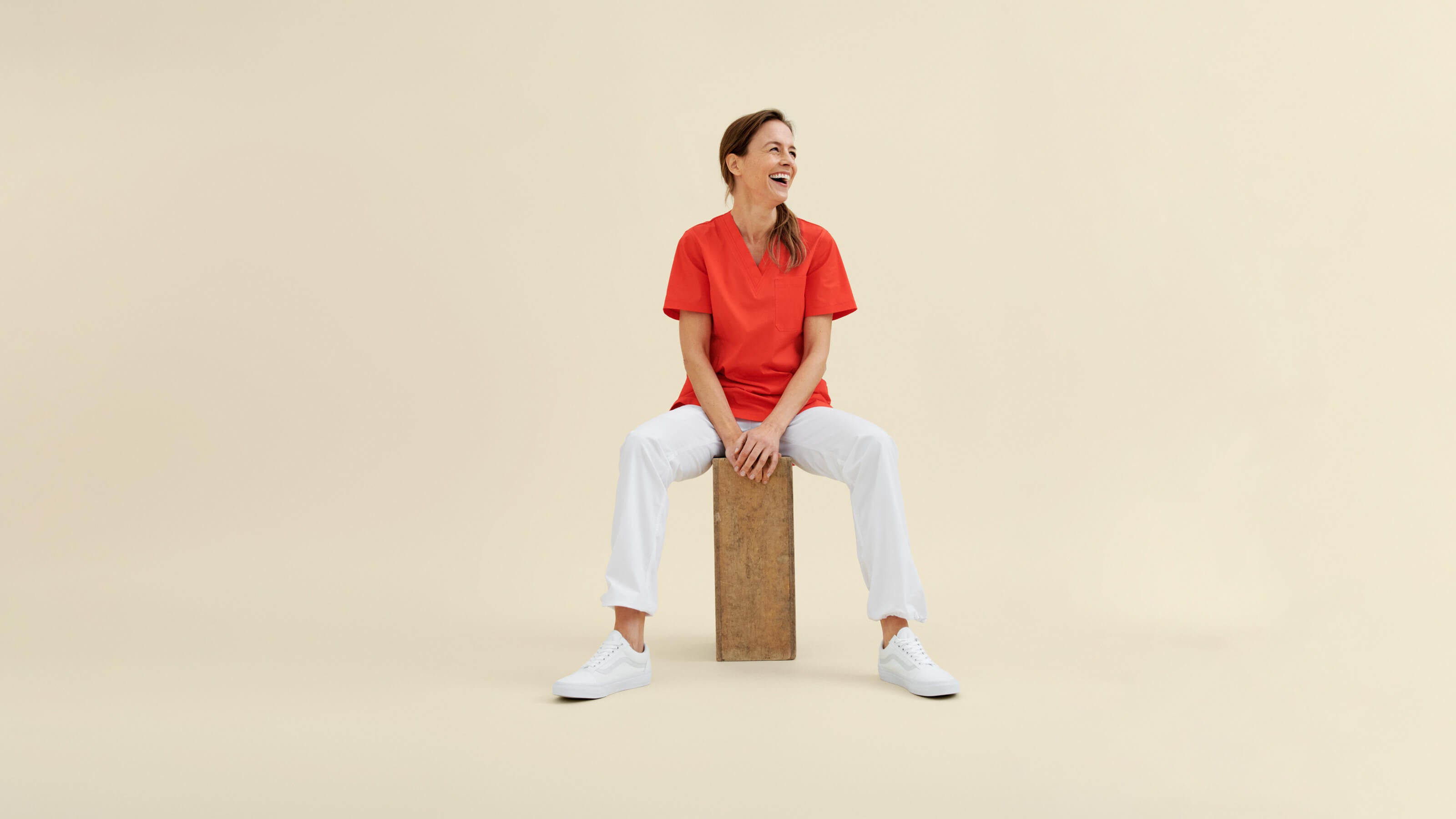 Hoitoalan punainen työpaita, valkoiset työhousut nauravan hoitajan yllä. Beige tausta.