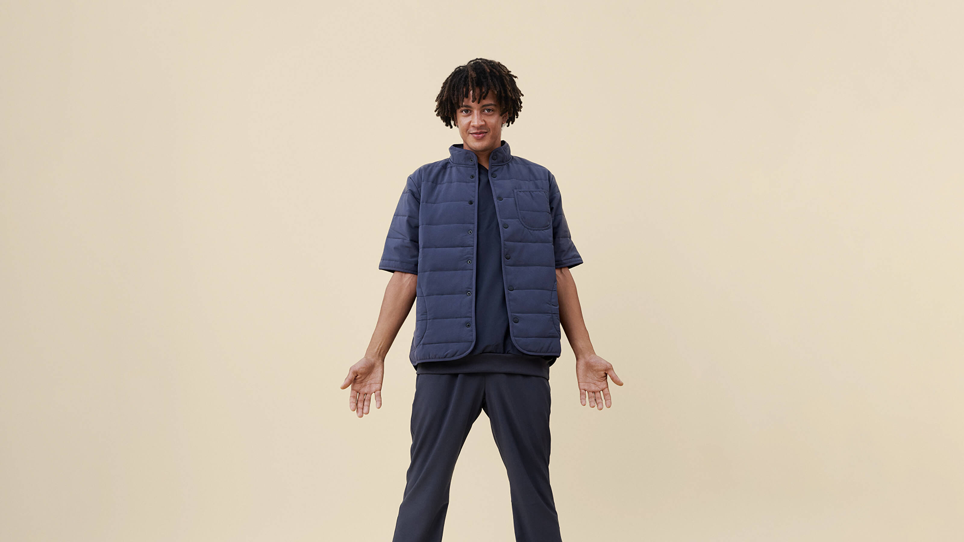 Tummansiniset vilutakki ja collegetyöasu seisovan, tummahiuksisen työntekijän päällä, beigellä taustalla