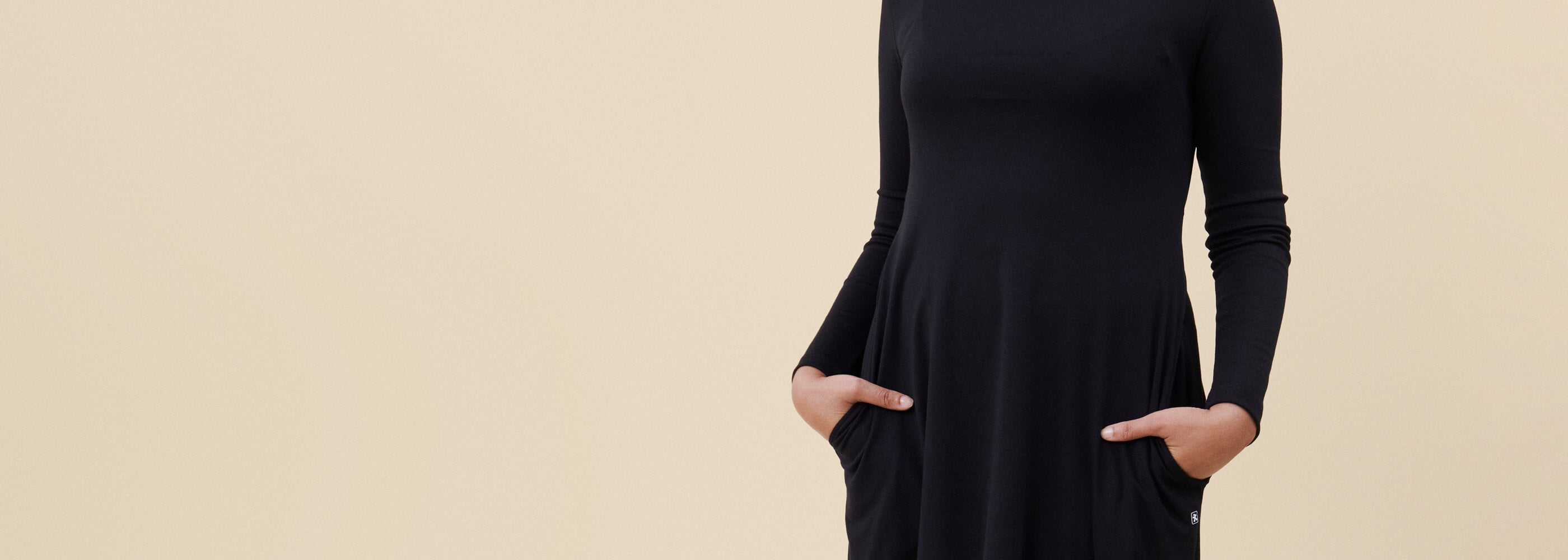 Musta työmekko työntekijän päällä, joustavaa materiaalia. Työntekijän kädet mekon taskuissa. Beige tausta.