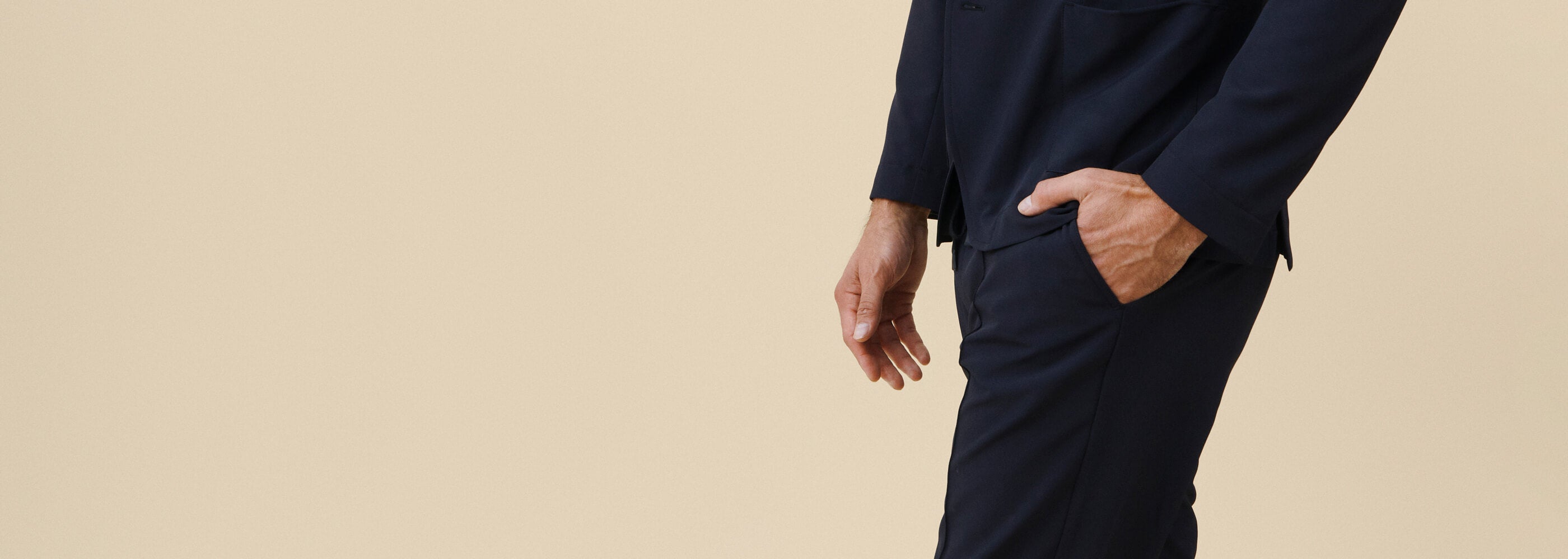 Tummansinen työasu toimistotyöntekijän päällä, sivusuunnasta. Mallin toinen käsi taskussa. Beige tausta.