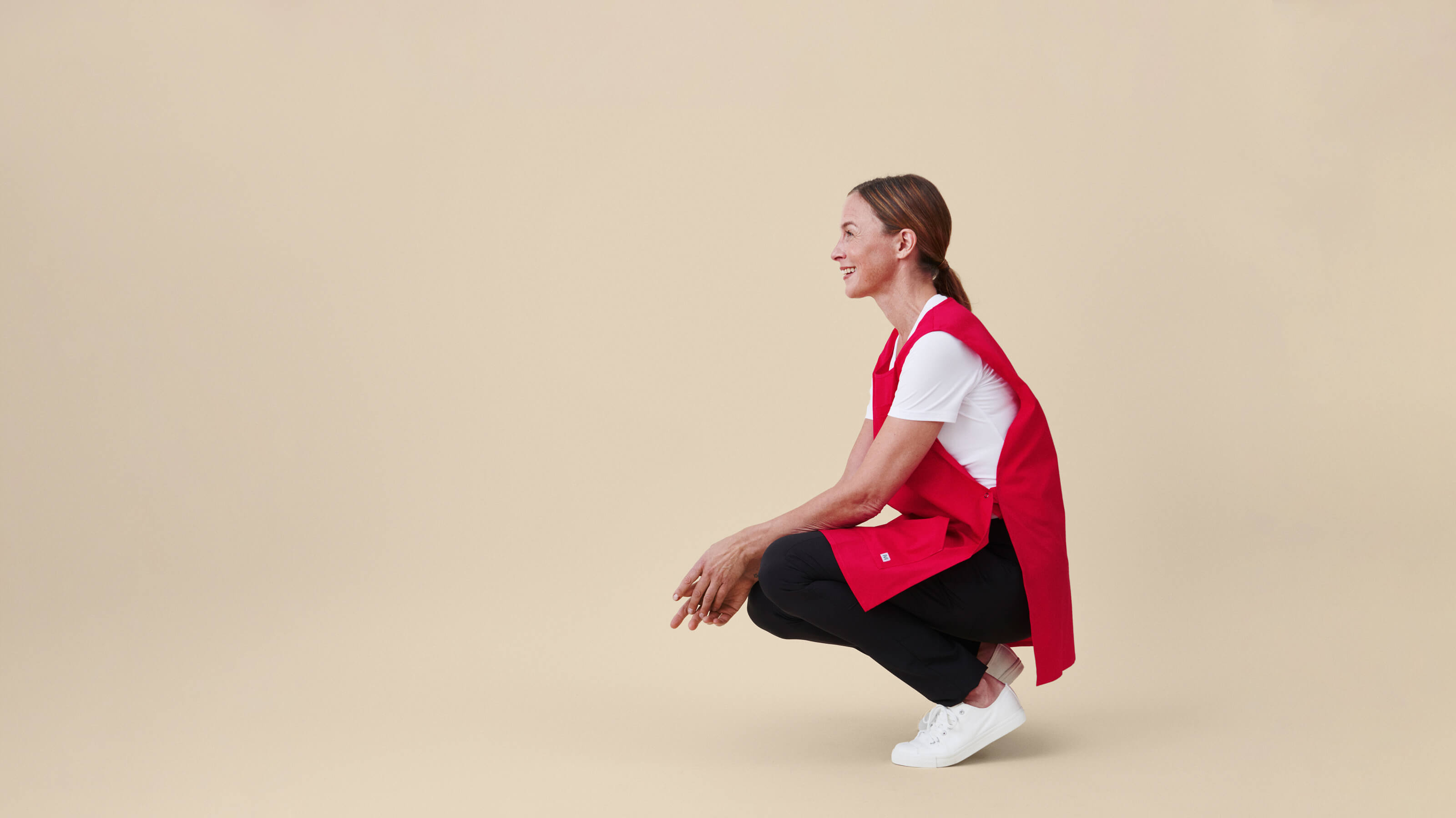 Punainen työessu, mustat työhousut ja valkoinen T-paita sivusuunnasta, hymyilevän hoitajan yllä. Beige tausta.