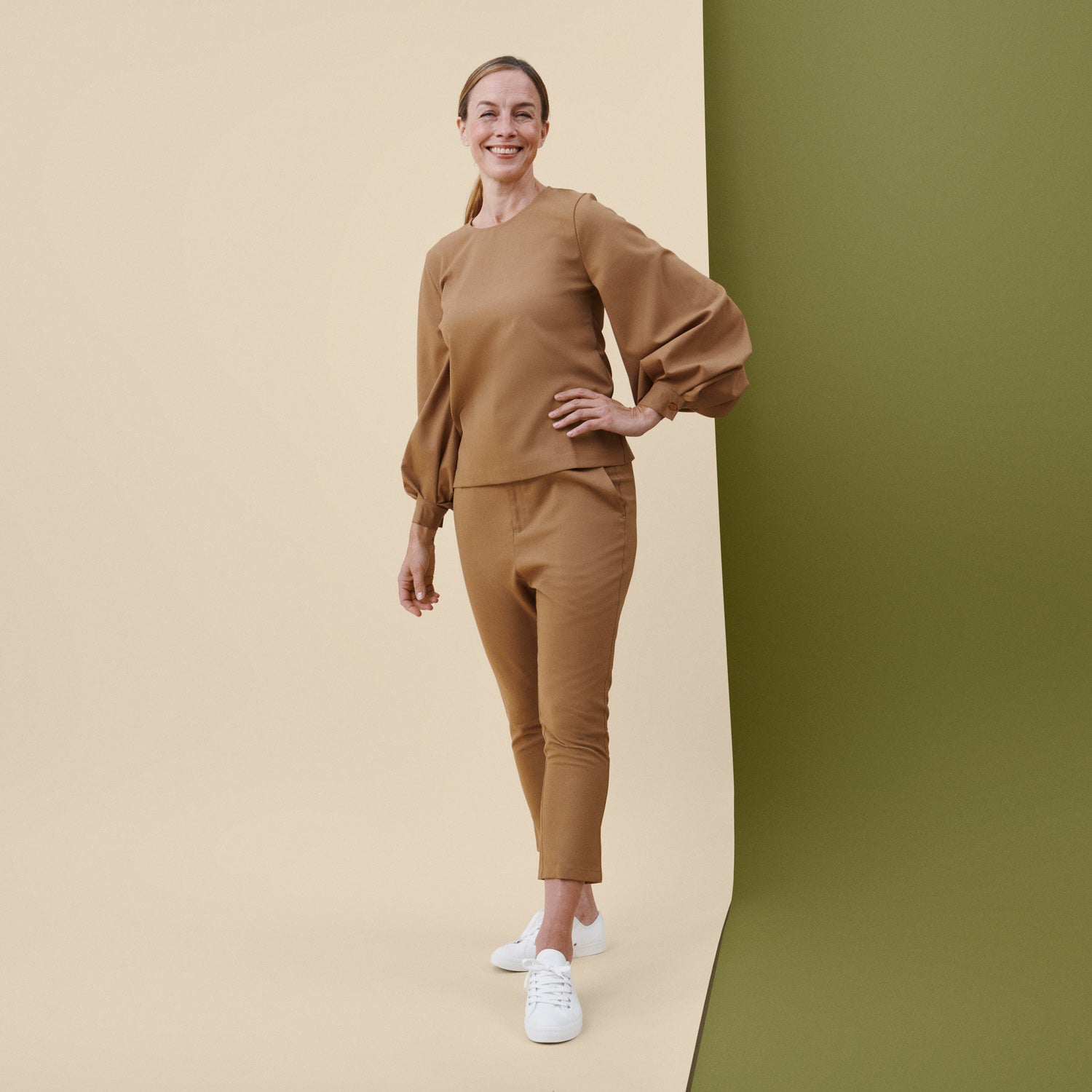 Vaaleanruskea työasu hymyilevän, seisovan työntekijän päällä, beigellä ja oliivinvihreällä taustalla