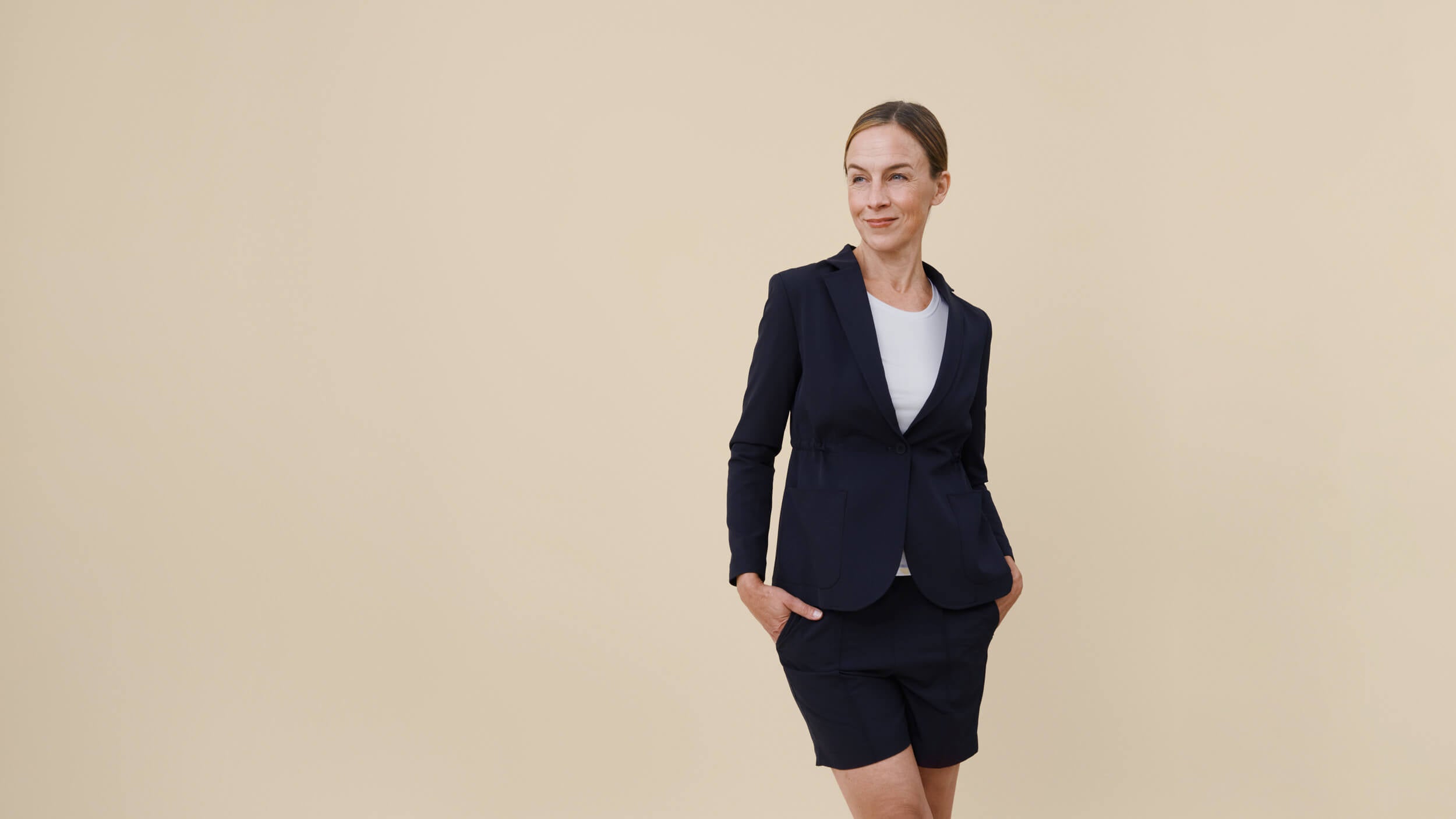 Tummansininen shortsi-bleiseri-asu sekä valkoinen työpaita seisovan toimistotyöntekijän päällä, beige tausta.