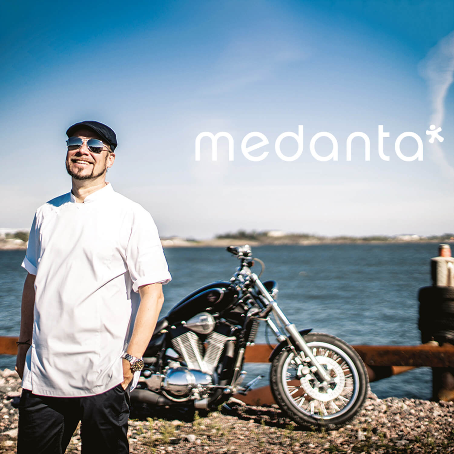 Medantan kokkimalliston työvaatteet hymyileväm kokin yllä. Taustalla merimaisema ja moottoripyörä.